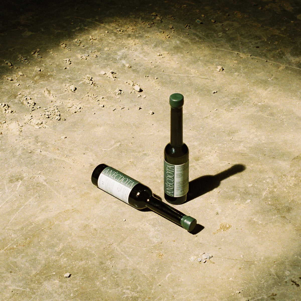 Dos botellas de aceite de oliva virgen extra Lagravera de Anècdota sobre un suelo de hormigón, una tumbada y otra de pie, con tapón de cierre y etiqueta en color verde que destacan en la composición.