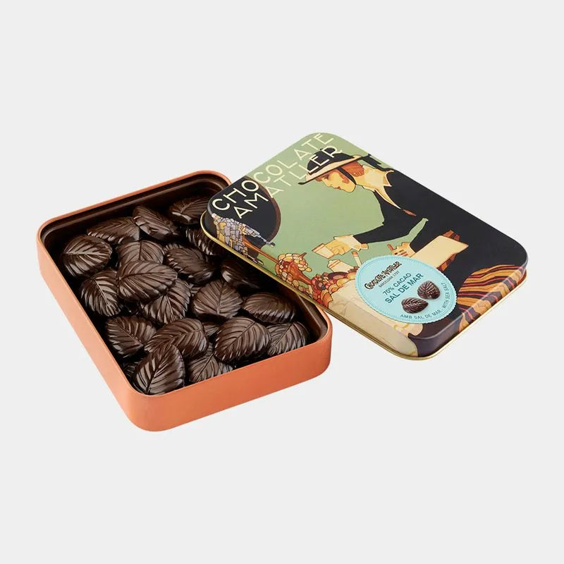 Lata metálica abierta con flores de chocolate negro con sal en su interior, sobre fondo de estudio. La tapa presenta una decoración inspirada en antiguos carteles de Amatller.