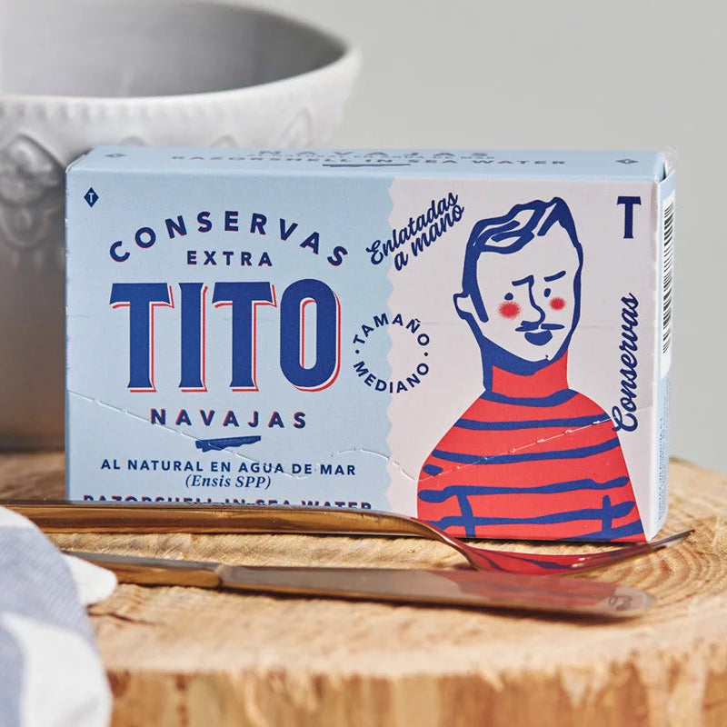 Lata de Navajas tamaño mediano de color azul claro y blanco con dibujo de marinero en un fondo de bodegón