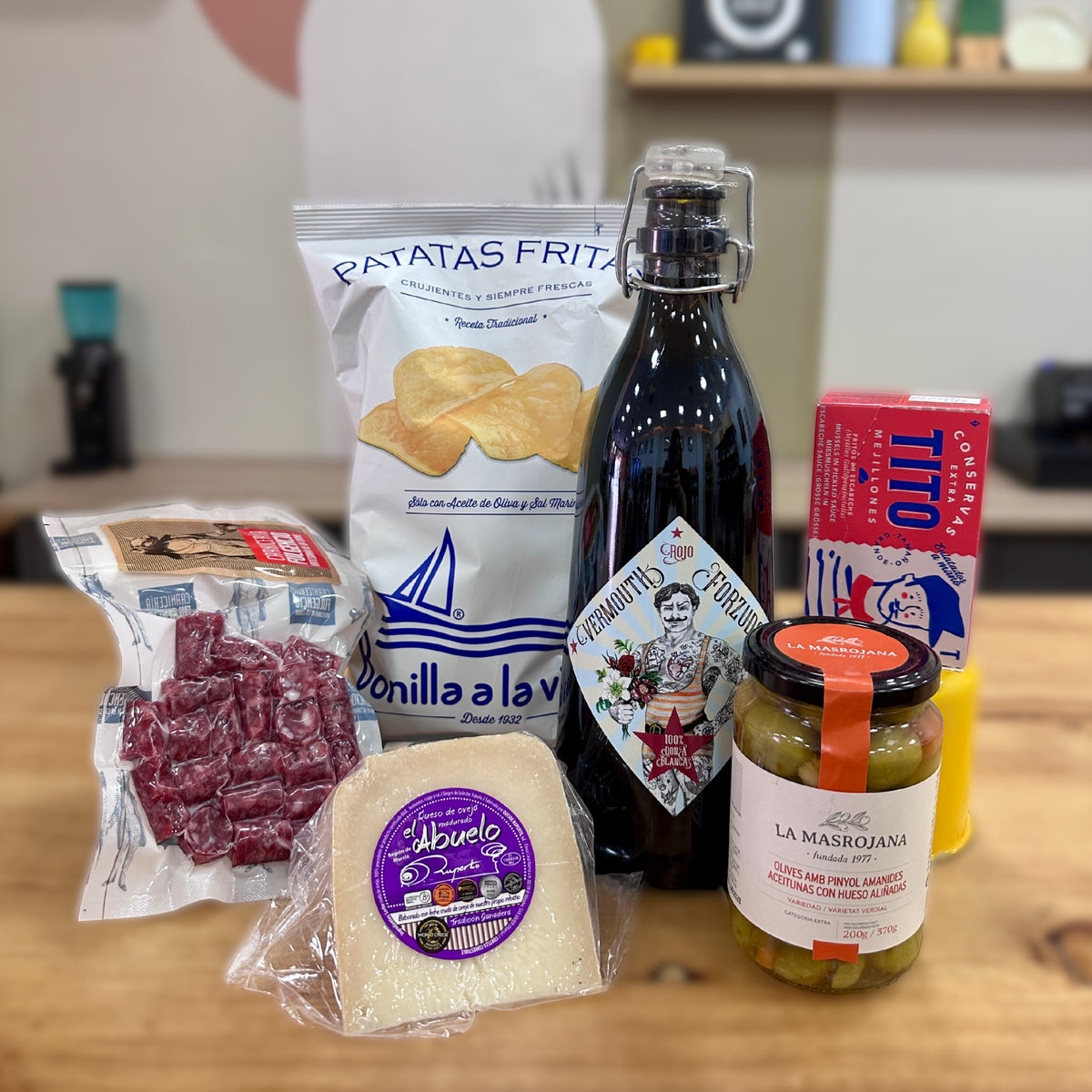 Pack Aperitivo Gourmet Clásico expuesto sobre el mostrador de la tienda D'12 Gourmet, compuesto por vermut rojo Forzudo, mejillones Tito, patatas Bonilla a la Vista, queso El Abuelo, aceitunas La Masrojana y salchicha de pellizco de chato murciano.