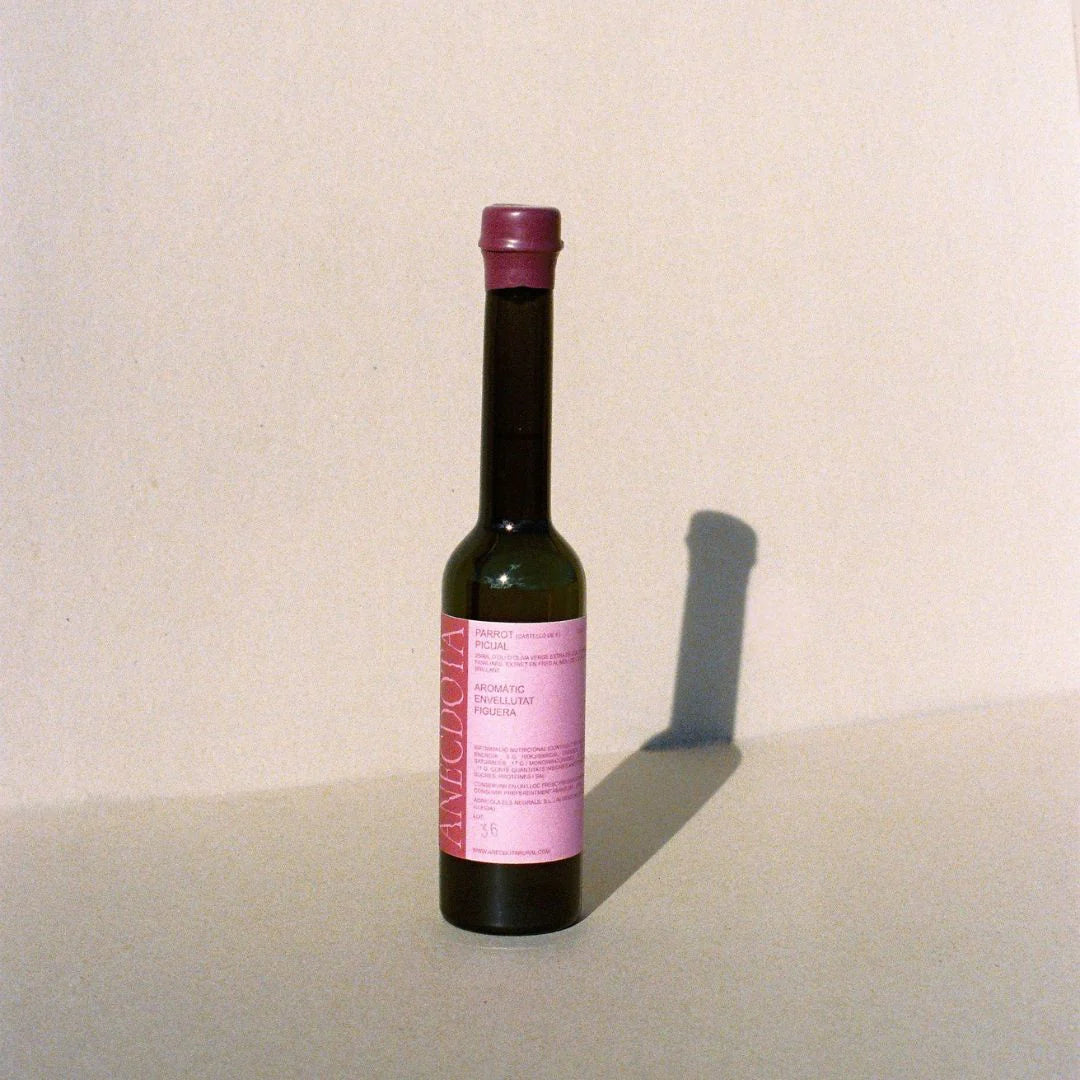Sobre fondo gris de estudio una botella de aceite de oliva virgen extra Parrot de la marca Anècdota, de la variedad Picual. Llama la atención los colores rosados tanto del tapón de cera como de la etiqueta.