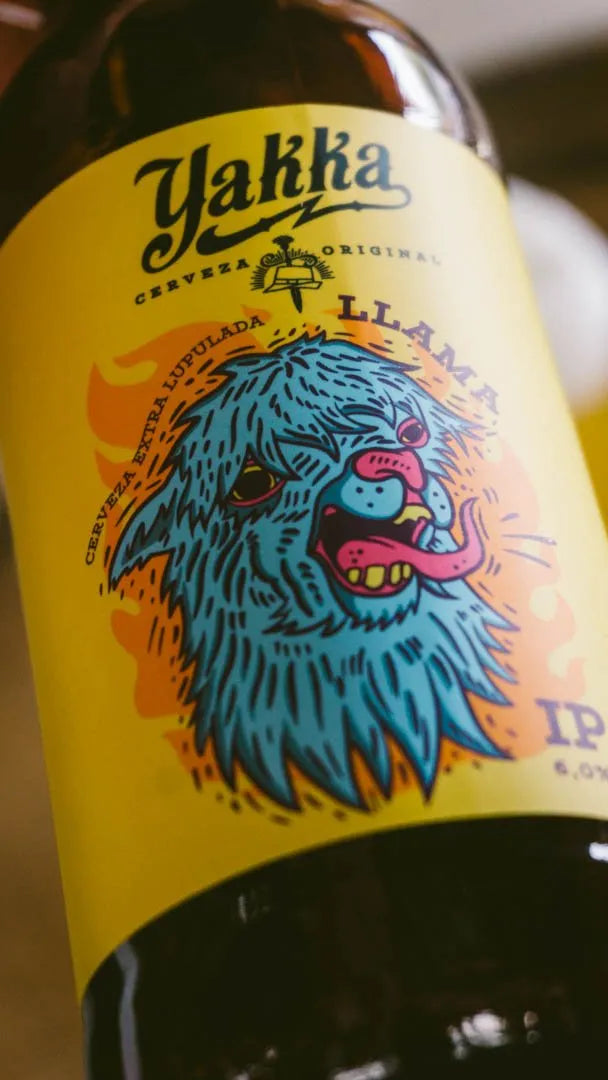 Primer plano de una botella de cerveza artesana Llama IPA de Yakka, colocada inclinada sobre un fondo de estudio, con la etiqueta amarilla y la ilustración azul de la cara de una llama claramente visibles.