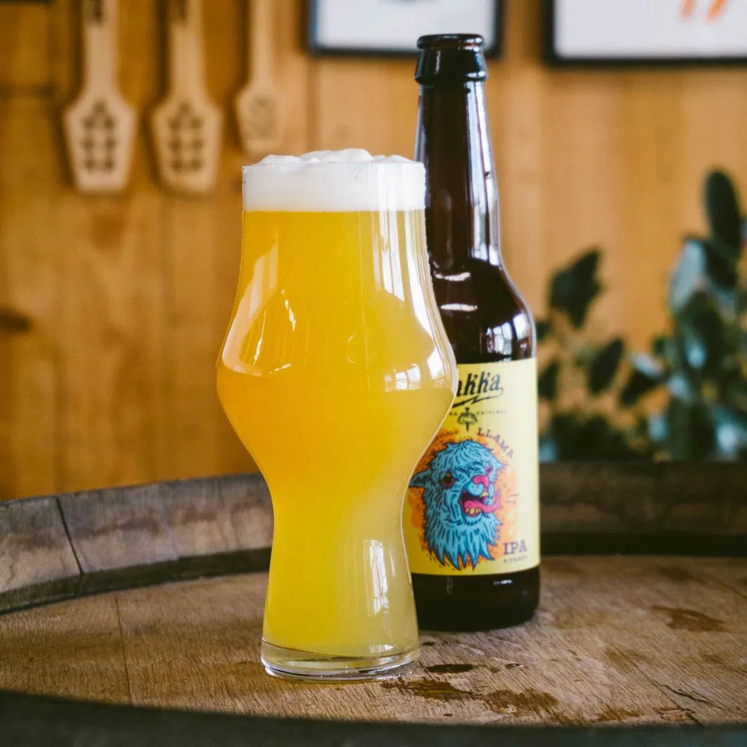 Vaso de cerveza artesana Llama IPA de Yakka sobre un barril de madera, con la botella de Llama parcialmente visible detrás. Al fondo se aprecia un bar desenfocado, creando una escena de ambiente cervecero.