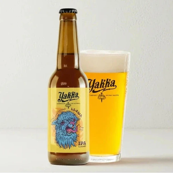 Botella de cerveza artesana Llama IPA de Yakka sobre fondo blanco de estudio, con etiqueta amarilla e ilustración en azul de la cara de una llama. Junto a la botella aparece un vaso servido con cerveza, donde se aprecia su color y la espuma en la parte superior.