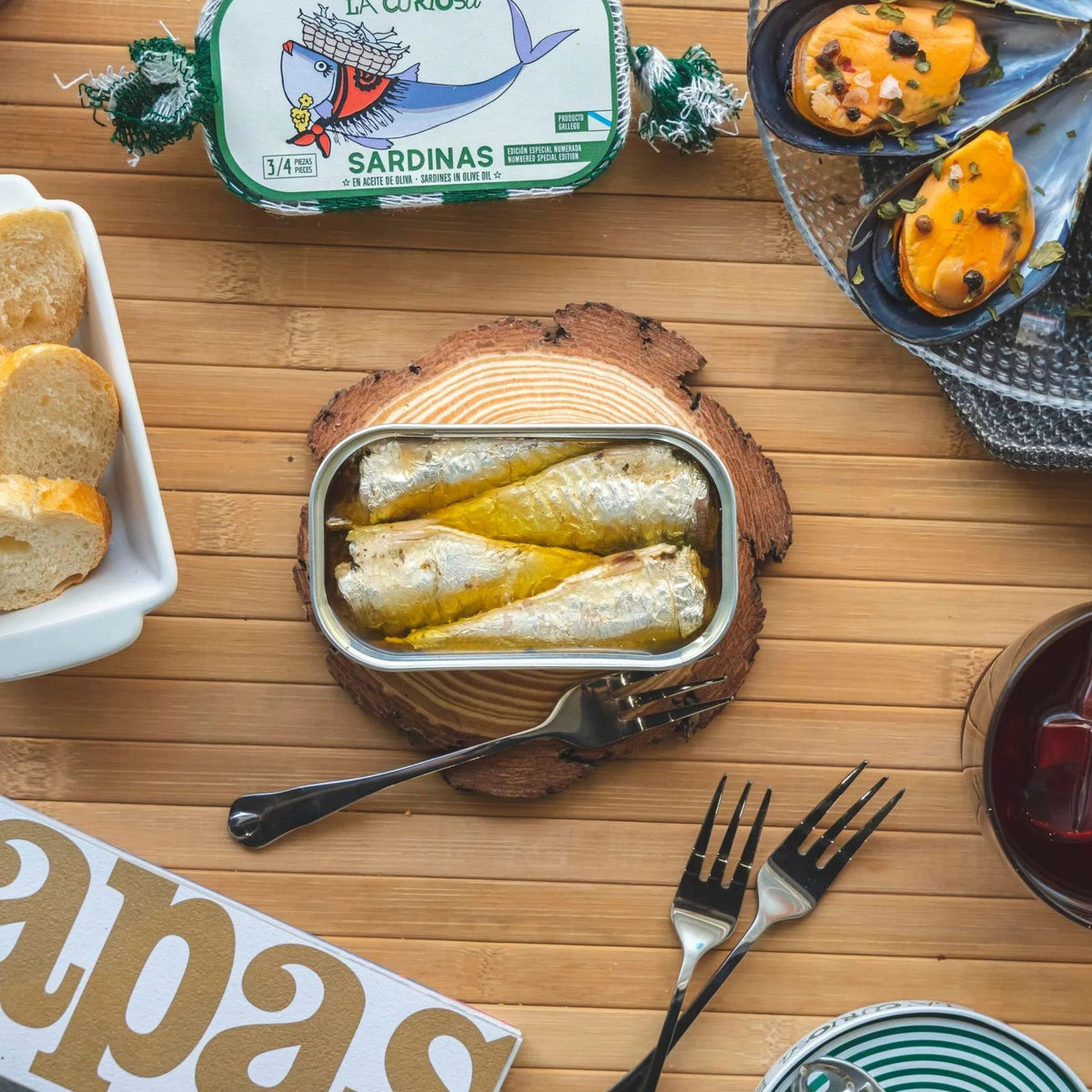 Mesa lista para el aperitivo con mejillones, pan y en el centro la lata abierta de sardinas La Curiosa.