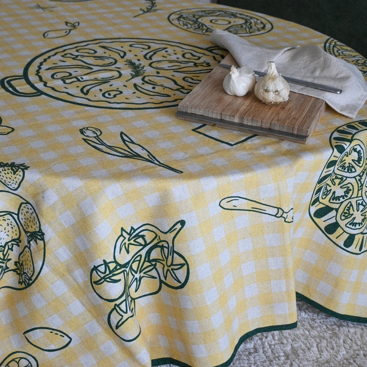 Una mesa redonda y sobre esta un mantel de rayas amarillas con motivos gastronómicos en verde. Sobre la mesa una tabla de madera con dos cabezas de ajos, cuchillo y trapo.