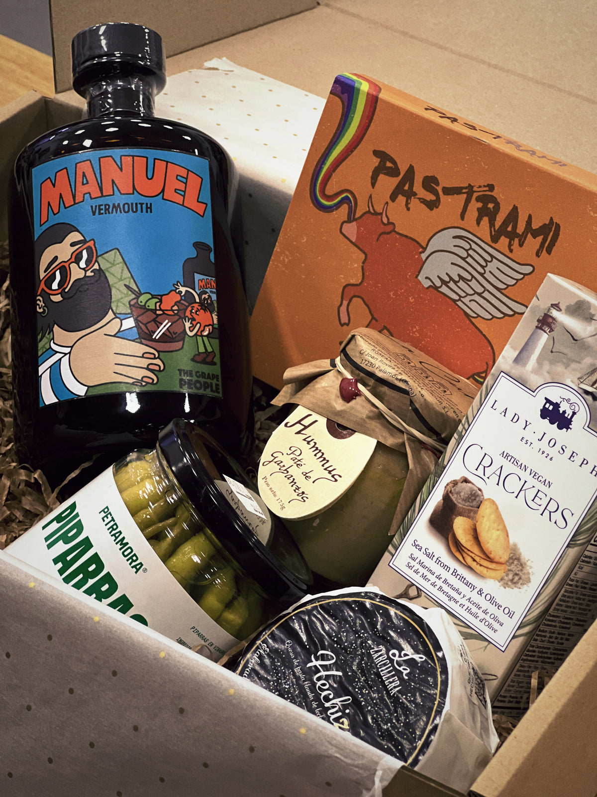 Caja Gourmet "Vermú, Pastrami & Rock’n Roll" en cartón característico de D'12 Gourmet, presentada con su contenido dispuesto en el interior: vermut Manuel, pastrami de Casa da Avoa, crackers saladas, piparras de La Gilda del Norte, hummus de La Cuna y queso Hechizo