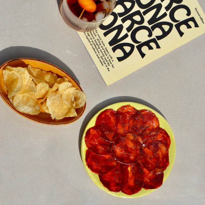 Mesa de aperitivo con un plato de chorizo ibérico de bellota loncheado de la marca LOLO, acompañado de un vaso de vermut y un plato de patatas.