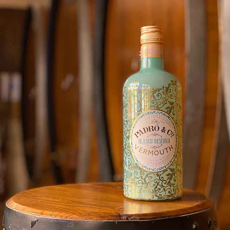 Botella de vermut blanco reserva de Padró & Co. botella elegante de color turquesa con toques dorados en forma de ramas. Sobre un barril de madera y el fondo se ve barriles.