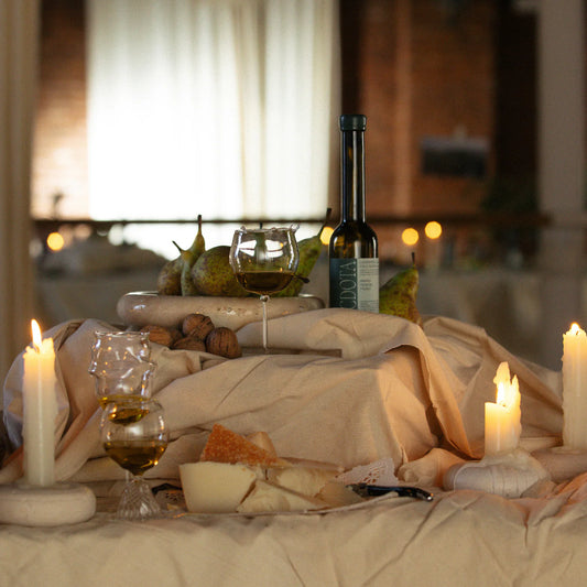 Vemos un fondo desenfocado parece ser un restaurante y en un primer plano una mesa con velas, vino, quesos, peras y el aceite Lagravera de Anécdota.