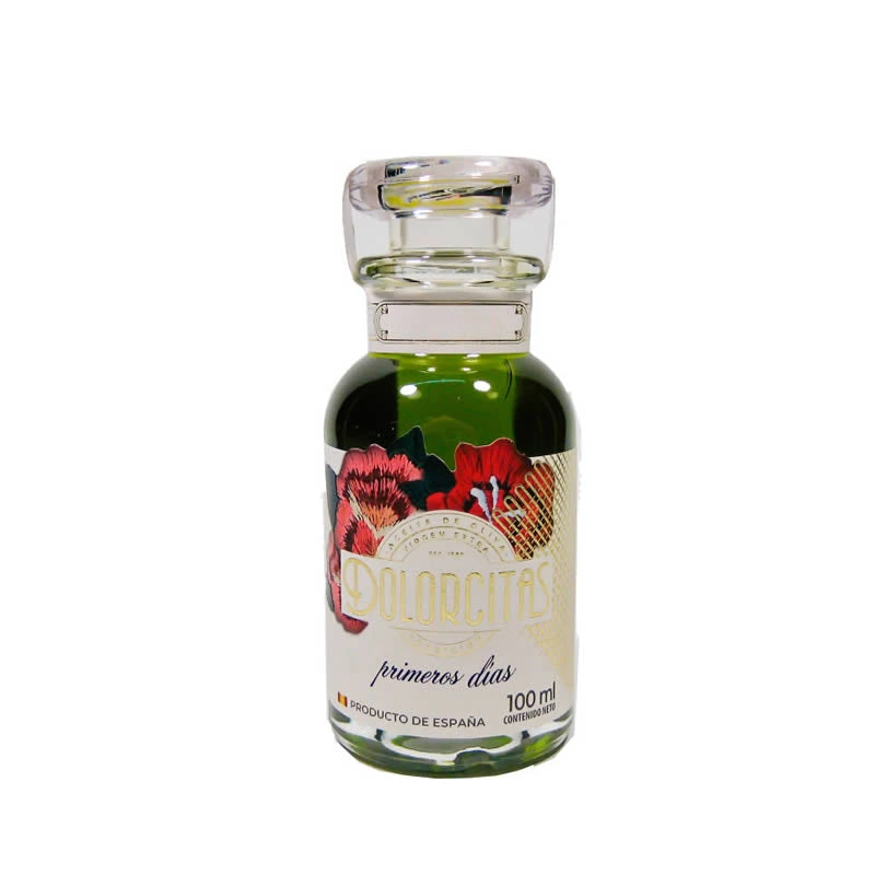 Fotografía de estudio sobre fondo neutro de la versión mini (100ml) en cristal transparente del aceite de oliva virgen extra Dolorcitas en D'12 Gourmet. Su pequeño envase permite apreciar la brillantez y el tono característico del aceite de alta calidad, luciendo una pulcra etiqueta blanca en el centro con un delicado motivo floral y el nombre de la marca en una tipografía sobria y elegante.