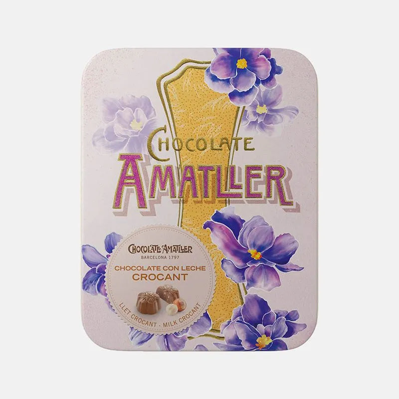 Lata metálica muy elegante con temática floral de tonos morados y amarillos. En su interior bombones en flor de chocolate con leche crocant de Chocolates Amatller.