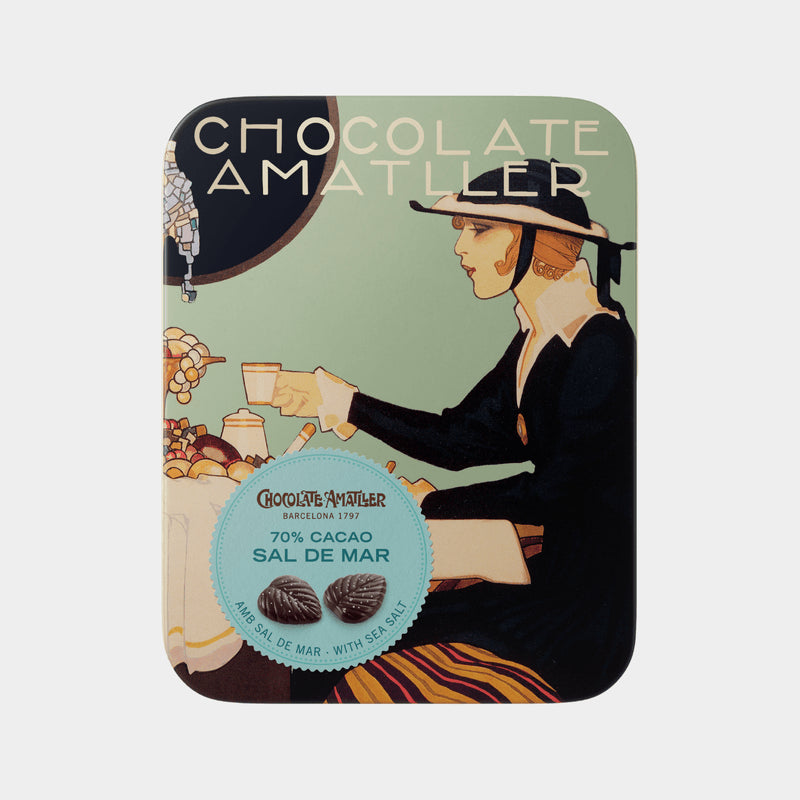 Lata metálica con flores de chocolate negro con sal de mar. La tapa de la lata se observa una bonita decoración de carteles antiguas de Chocolates Amatller