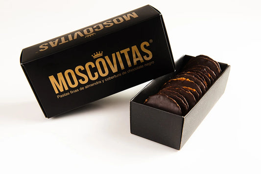 Caja abierta de Moscovitas Dark mostrando las finas pastas de almendra con cobertura de chocolate en su interior, junto a una caja cerrada de color negro con el nombre MOSCOVITAS en grandes letras doradas.