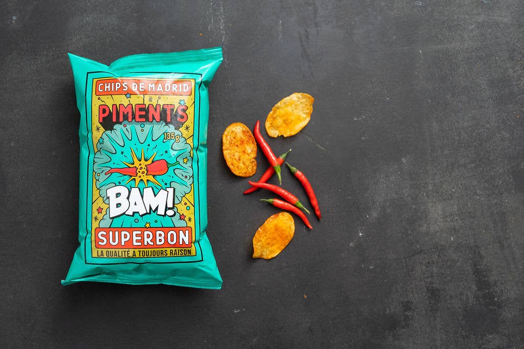 Bolsa de patatas sabor chili Superbon sobre fondo negro tipo pizarra, con envase azul de tipografía moderna y llamativa en varios colores, acompañada de tres patatas y tres chilis.