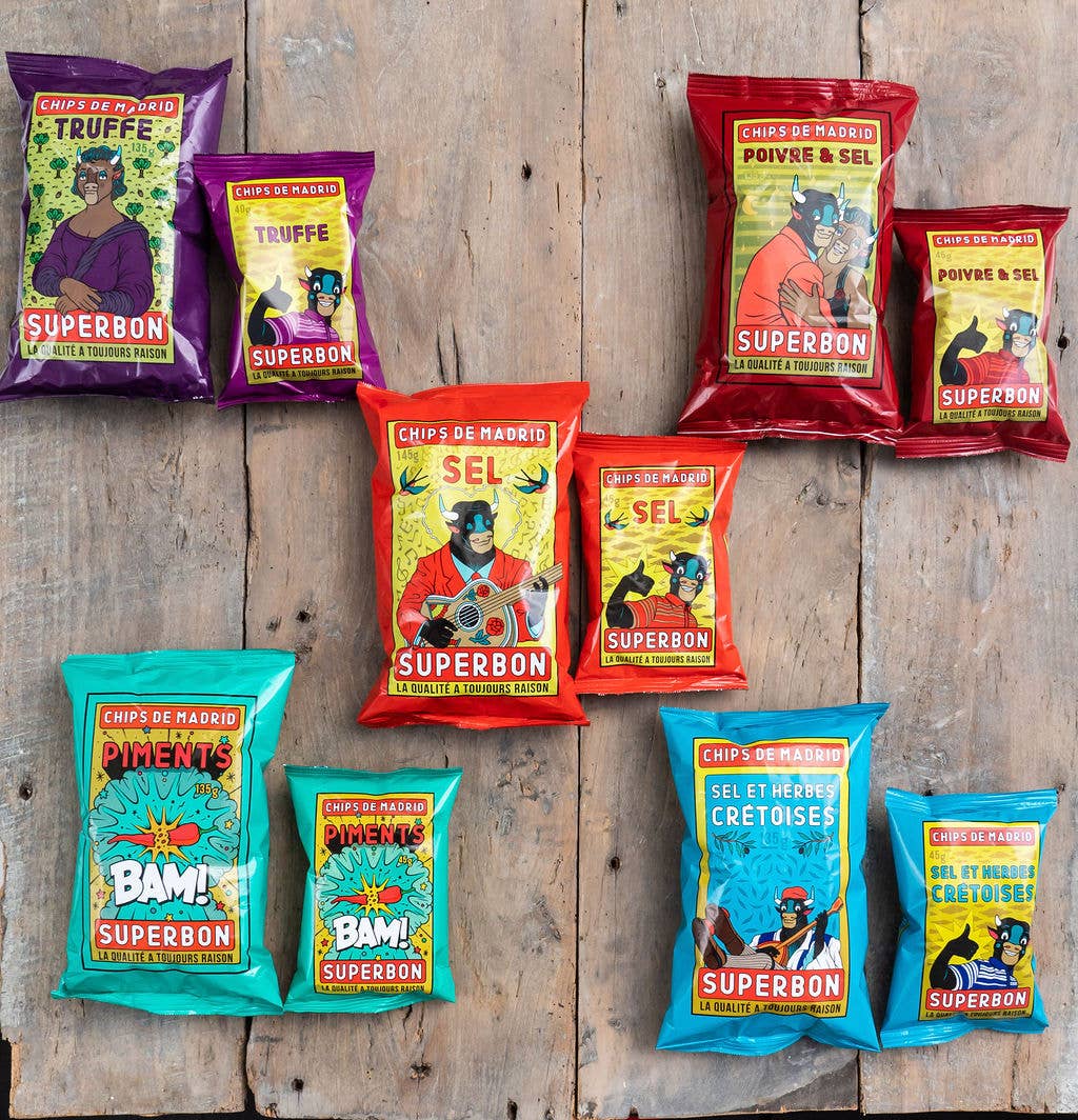 Selección de patatas fritas Superbon sobre una mesa de madera con los sabores trufa, sal marina, chili y hierbas de Creta en formatos grande y pequeño. Los envases presentan un diseño moderno con ilustraciones de una vaca humanizada, tipografía llamativa y colores vivos como azul, rojo, morado y verde.
