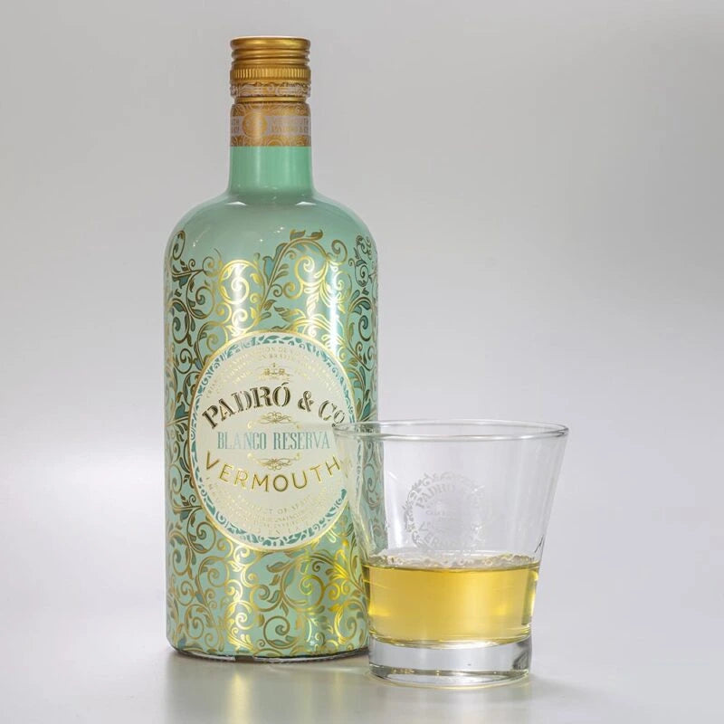 Botella de vermut blanco reserva de Padró & Co. botella elegante de color turquesa con toques dorados en forma de ramas. Acompañado de un vaso de vermut donde podemos ver el tono y la calidad de este vermut.