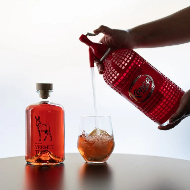 Botella de Vermut Tres Rucio junto a un vaso lleno de vermut, el cual se está llenando gracias a un sifón rojo.