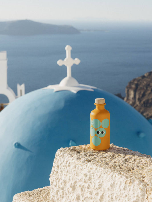 Botella de vinagre balsámico blanco Yiayia & Friends anaranjada con lunares turquesa y tapón de madera sobre un muro de piedra blanca, con una cúpula azul de estilo griego al fondo en D'12 Gourmet