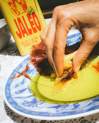 Botella de aceite de oliva virgen extra Jaleo sobre una mesa junto a un plato con aceite mientras una mano moja pan, recreando una escena de degustación y aperitivo.