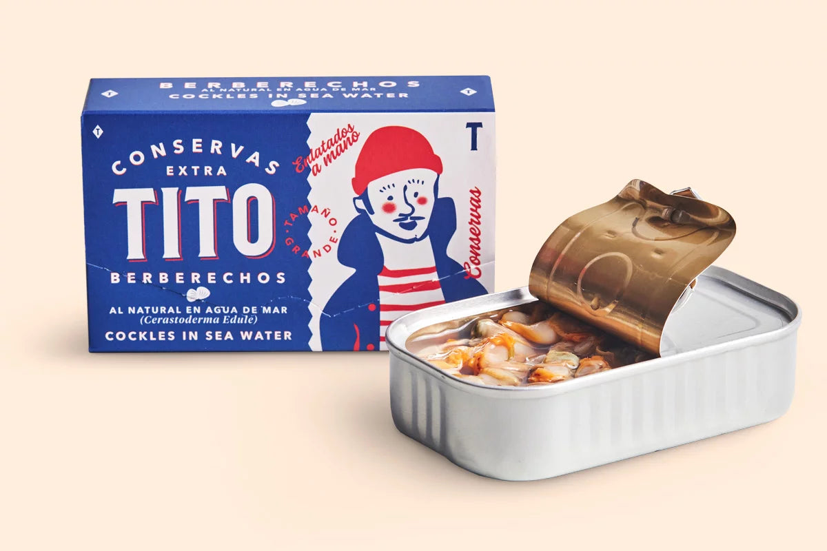 Lata de conservas de Berberechos al natural de Conservas Tito. Lata azul con letras blancas y dibujo de marinero, junto a esta una lata de berberechos semi-abierta.