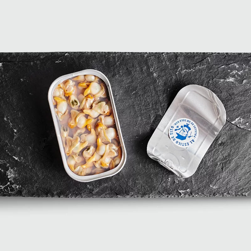 Sobre plato de pizarra una lata de conservas de berberechos al natural abiertas de Conservas Tito.