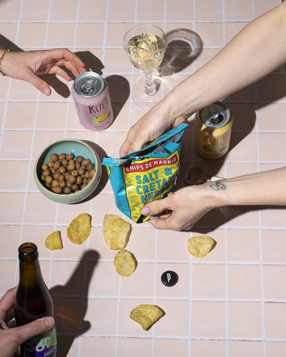 Vemos una mesa con aceitunas, vino y unas manos abriendo una bolsa de patatas de sal y hierbas de creta de Superbón.