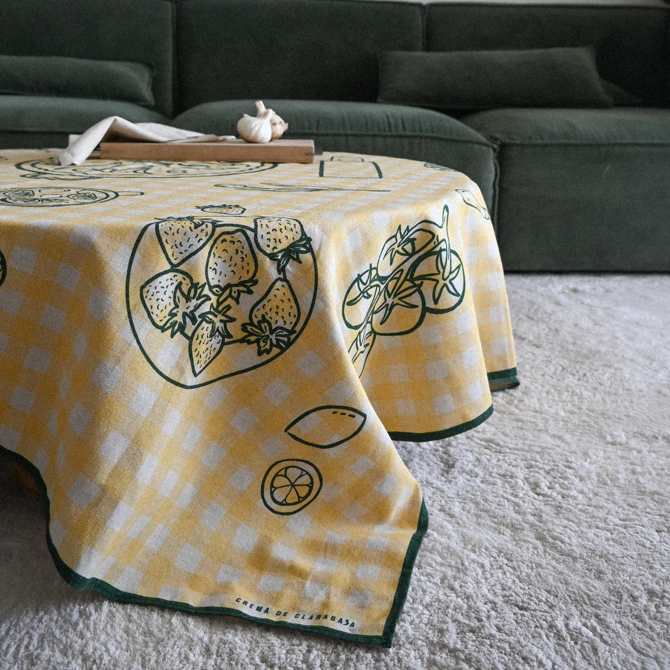 Mantel Vichy amarillos con motivos gastronómicos en verde sobre una mesa redonda. En un salón, donde vemos en el fondo en un salón con sofá verde y en el suelo una alfombra gris.