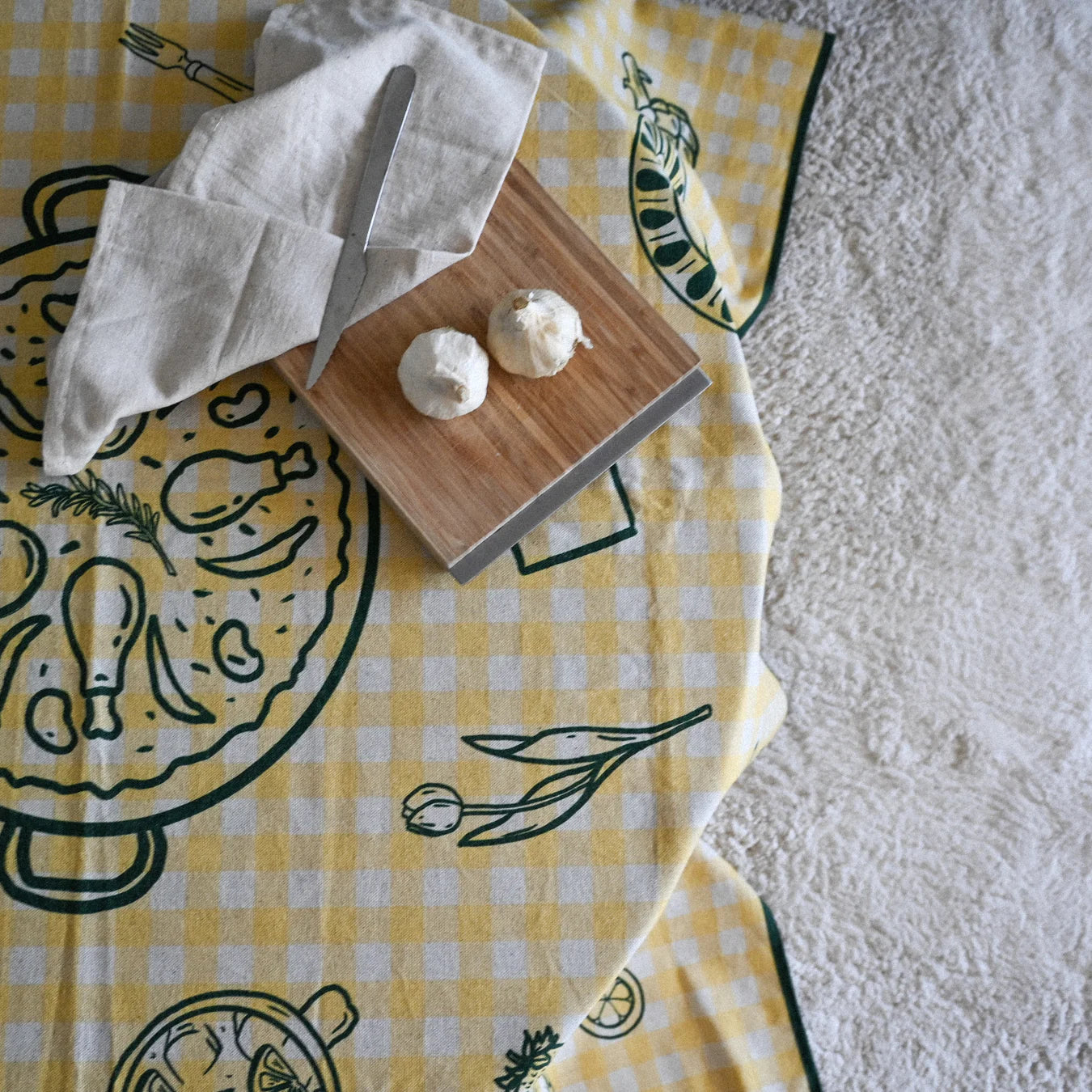 Mesa redonda con mantel a rayas amarillo con motivos gastronómicos en verde. Sobre la mesa una tabla con dos cabezas de ajo, cuchillo y trapo. en el suelo tiene una moqueta gris.