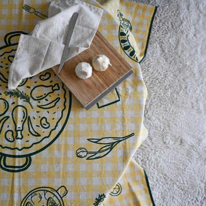 Mesa redonda con mantel a rayas amarillo con motivos gastronómicos en verde. Sobre la mesa una tabla con dos cabezas de ajo, cuchillo y trapo. en el suelo tiene una moqueta gris.