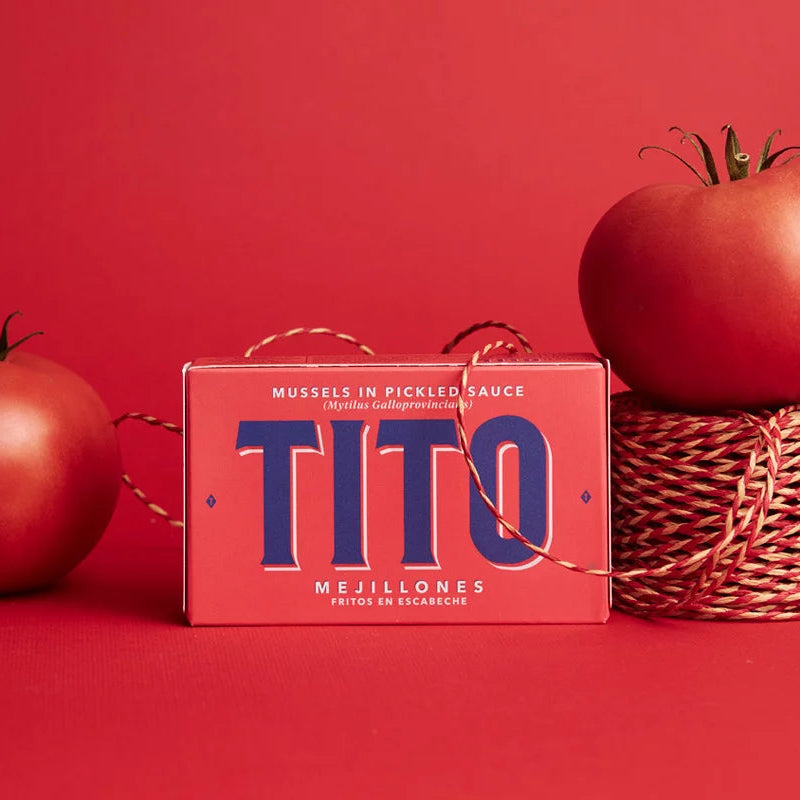 Bodegón con tomates y fondo rojo, en el centro encontramos una lata de Mejillones fritos en escabeche de color rojo con letras azules.