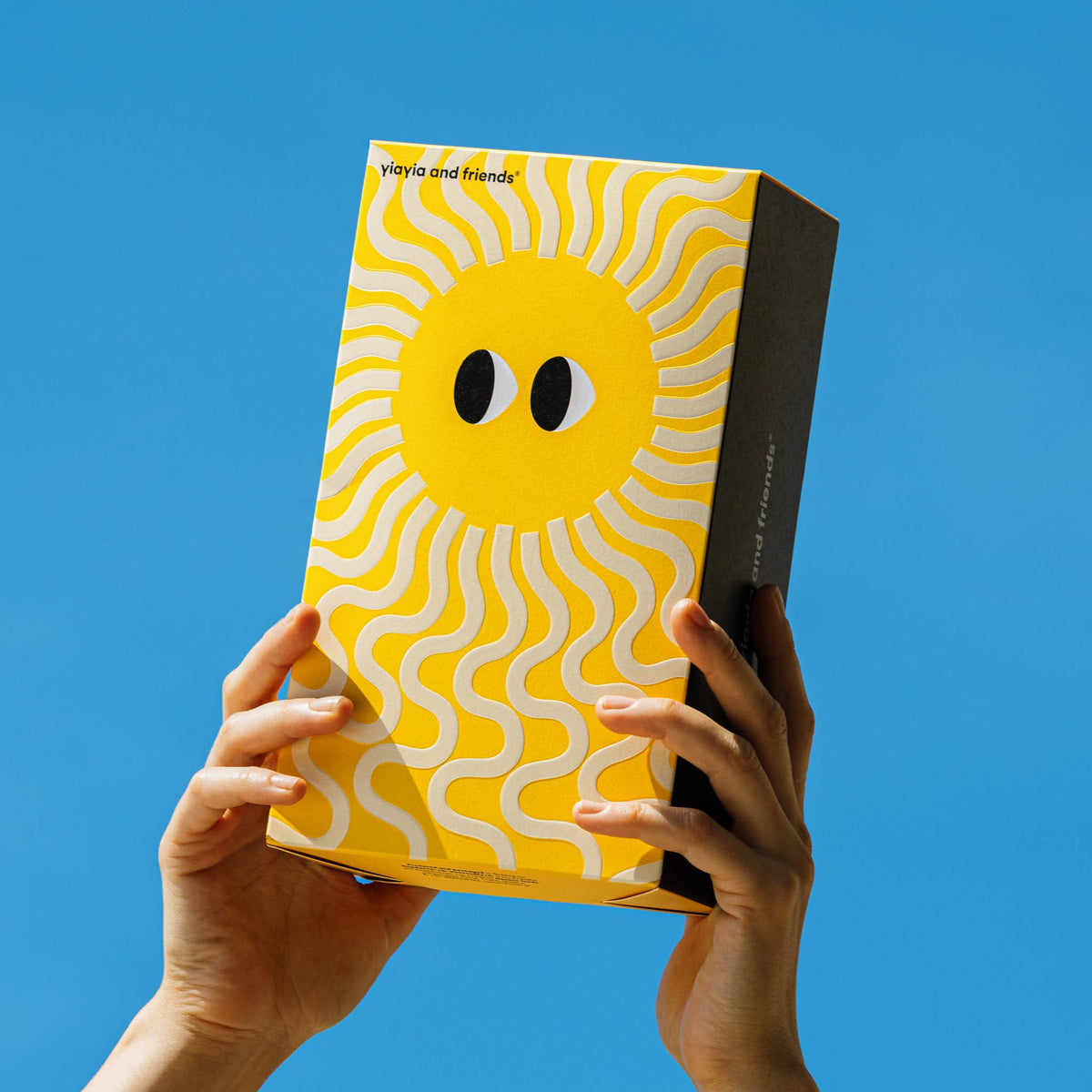 Caja de pasta griega Hylopites de Creta Yiayia & Friends alzada por dos manos con fondo de cielo azul, envase amarillo e ilustración del sol con ojos de Yiayia en D'12 Gourmet