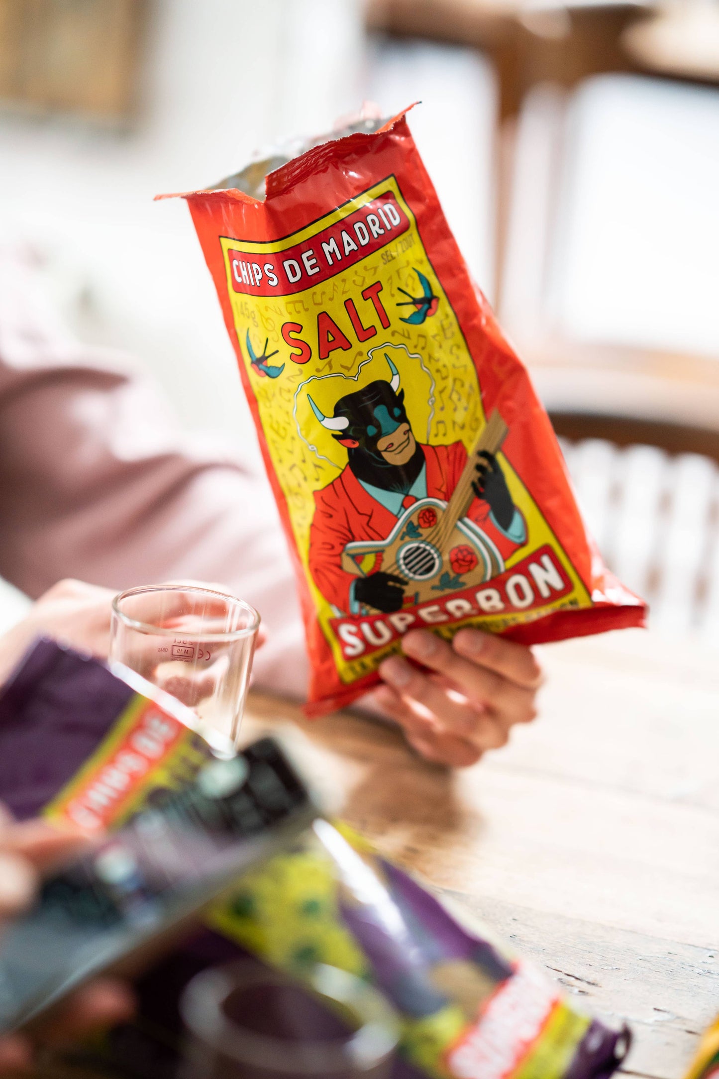 Una persona sostiene una bolsa de patatas fritas de sal marina Superbon, con envase de color rojo e ilustración central de una vaca humanizada tocando una guitarra.