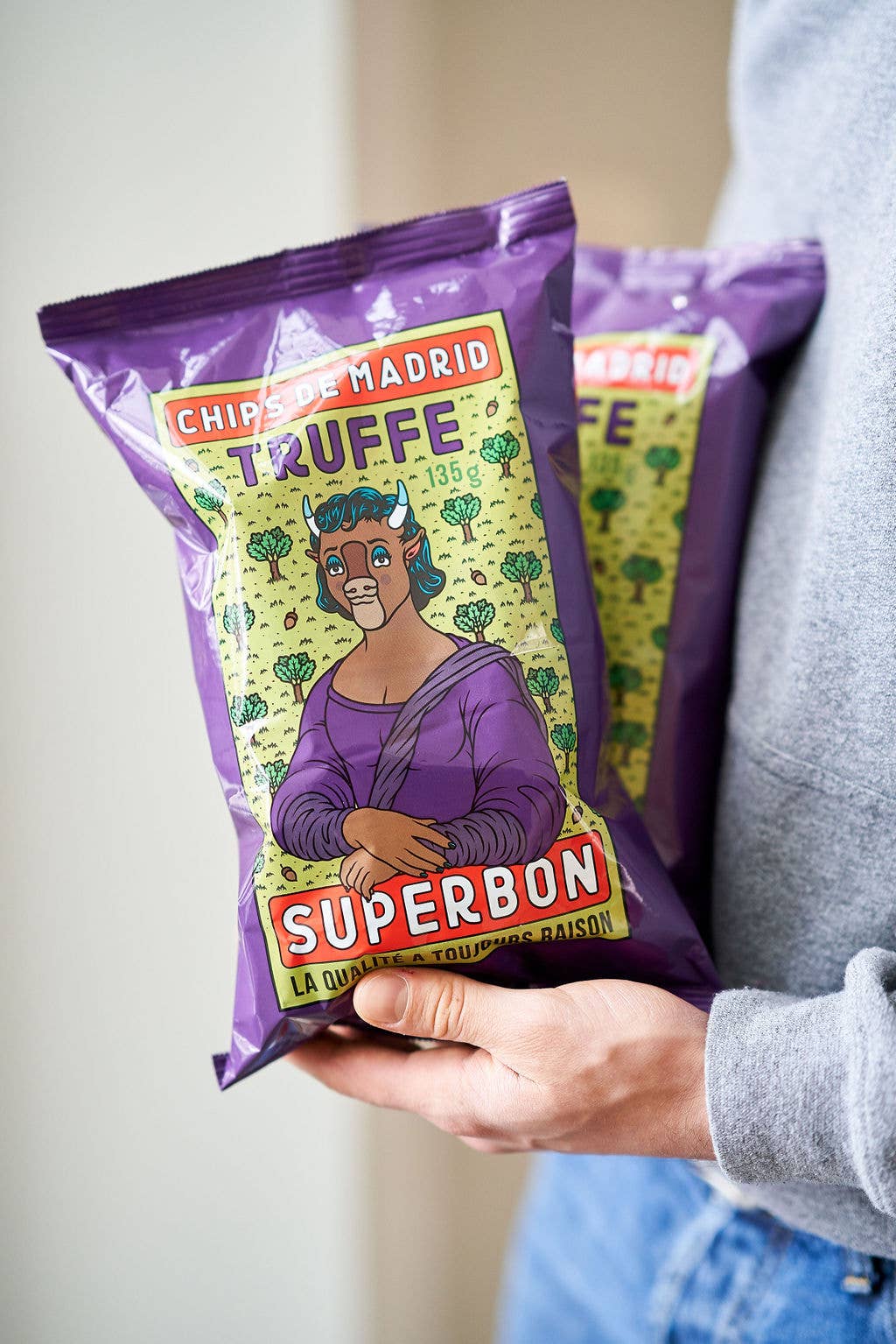 Una persona sostiene dos bolsas de patatas fritas sabor trufa Superbon, con envases de color morado e ilustración central de una vaca humanizada inspirada en la Mona Lisa.