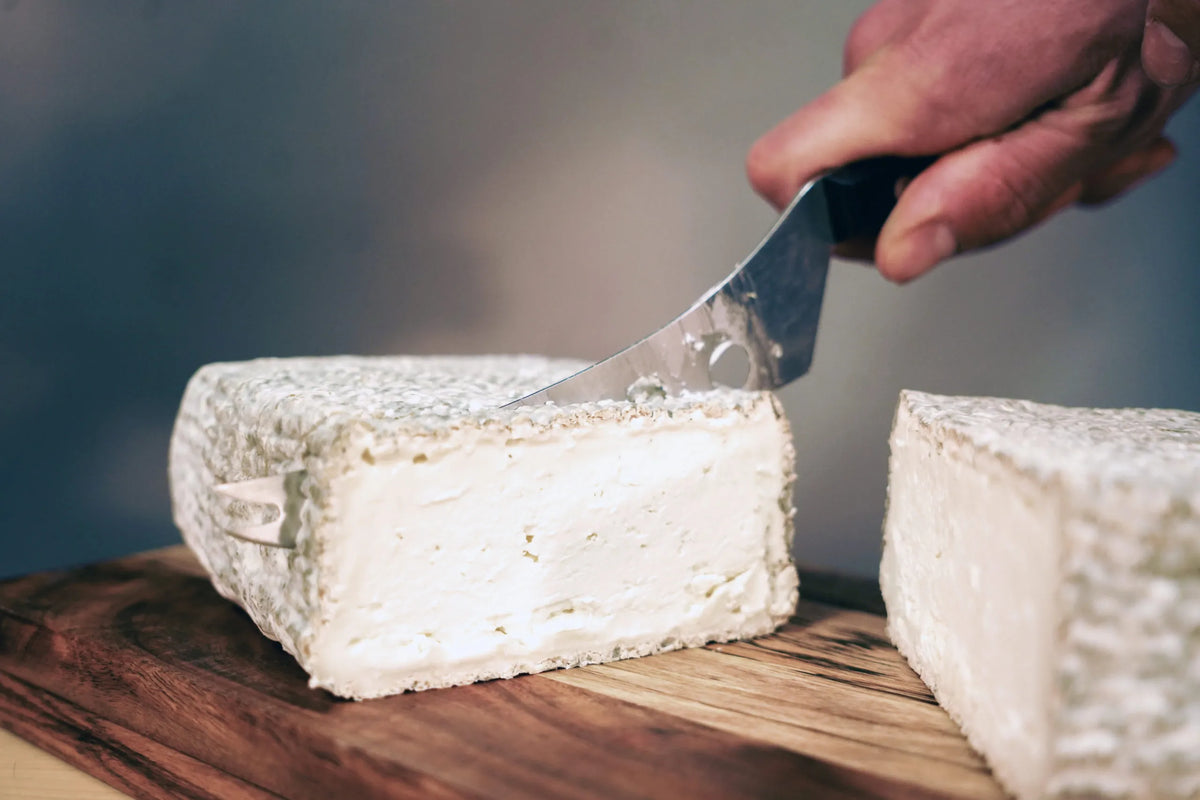 Mano cortando por la mitad una pieza entera de queso de cabra El Hechizo de la Zarcillera, mostrando su corteza exterior y su interior blanco y cremoso - D'12 Gourmet
