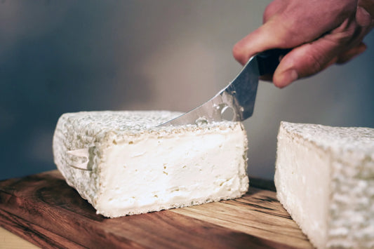 Mano cortando por la mitad una pieza entera de queso de cabra El Hechizo de la Zarcillera, mostrando su corteza exterior y su interior blanco y cremoso - D'12 Gourmet