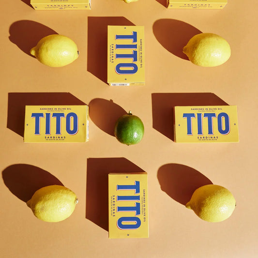Latas de Sardinas pequeñas de Tito, de color amarillo y letras azules. Junto a limones y una lima