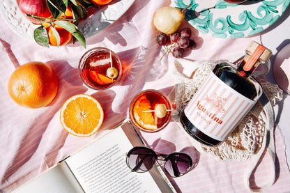 Mesa con libro abierto, gafas de sol, naranjas y dos vasos con vermut Agustina. También vemos la botella de Vermut Agustina.