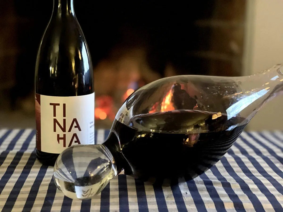 Botella de vino tinto Tinaja junto a un decantador que muestra el color del vino, sobre una mesa con mantel de cuadros azules y una chimenea tradicional al fondo.