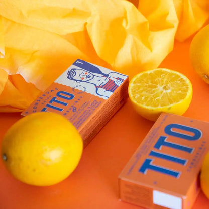 Latas naranjas con zamburiñas en salsa de vieiras de Conservas Tito entre limones y sobre una mesa naranja.